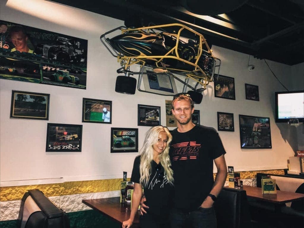 A race car and his blonde girls at an eatery where on of his old race cars are displayed.