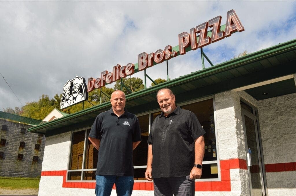 A photo of two men in front of a pizza shop.